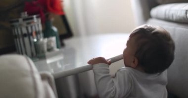 Baby standing up holding into table furniture in living-room with hand, cute infant learning to stand up