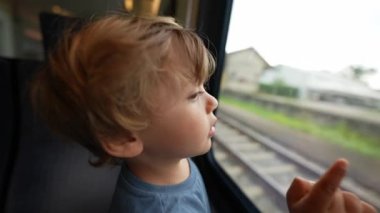 Child travels by train leaning on window pointing at landscape passing by