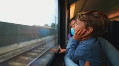 Anne ameliyat maskesi takıyor. Trenin içinde çocuk tutuyor.