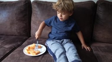 Adorable child eating melon fruit sitting on couch small boy eats healthy snack fruit with fork