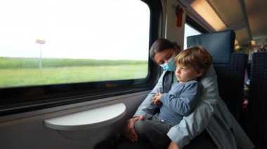 Mother holding toddler in arms traveling by train wearing face mask
