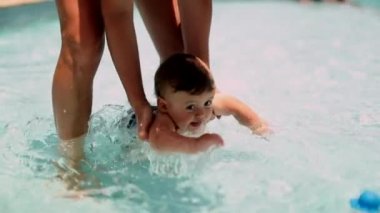 Happy baby at the swimming pool splashing water. Mother helping toddler infant splashes water