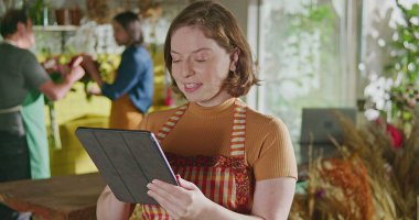Happy female staff of flower shop holding tablet checking online orders. Young woman employee using modern technology to manage shipment delivery of small business store