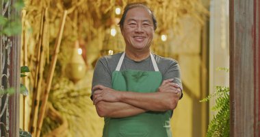 An Asian American business owner wearing apron and crossing arms smiling. Portrait of an asian middle aged Businessman owner of small local store with arms crossed looking at camera