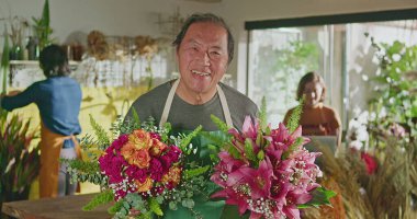 Asian American business entrepreneur owner of flower shop standing holding two bouquet of flower arrangements smiling at camera. Small business concept of floriculture store with employee working in the background