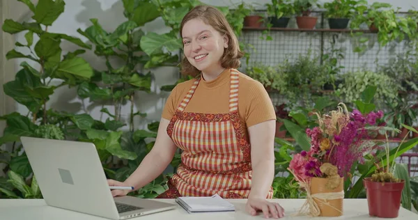 Portrait of a female employee wearing apron in front of laptop smiling at camera receiving online orders at small business flower shop retail store. Happy worker in front of computer