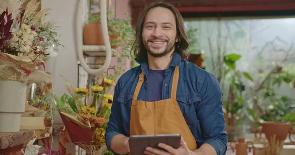 Portrait of a young small business entrepreneur holding tablet and wearing apron inside shop smiling at camera