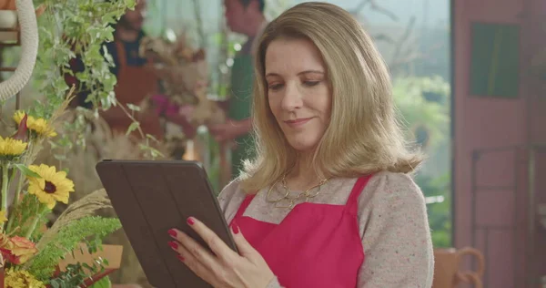 Female business owner of retail store using tablet technology to check inventory while employees work in the background. A middle aged business woman entrepreneur owner of small business store