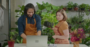 Young entrepreneurs managing online orders at small business flower shop. Male a female employee staff wearing apron writing notes using internet to expand business