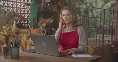 Owner of small business taking online orders in front of laptop computer. Female entrepreneur of local flower shop talking with customer or partner online wearing apron talking on phone