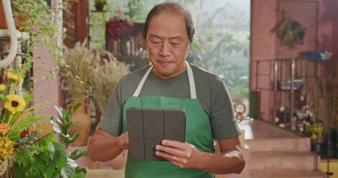 Portrait of a happy Asian American business owner holding tablet smiling at online orders. Male entrepreneur of small business store using modern tablet digital device to manage inventory
