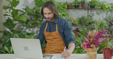 Young florist entrepreneur checking online orders smiling in front of laptop. Small business online orders concept. Male employee wearing apron writing down online orders
