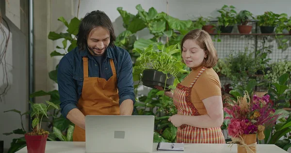 Young entrepreneurs managing online orders at small business flower shop. Male a female employee staff wearing apron writing notes using internet to expand business