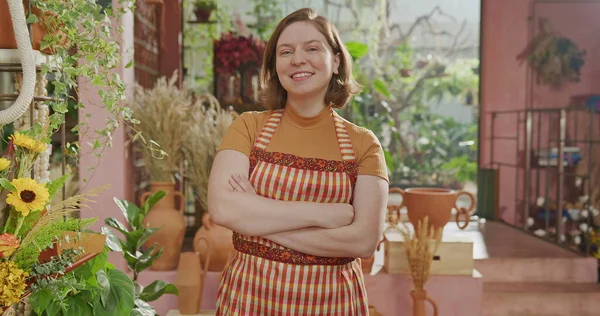 Portrait of female staff wearing apron standing inside small business smiling at camera. Happy worker wearing apron looking at camera inside flower shop