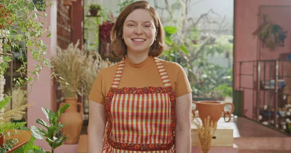 Portrait of female staff wearing apron standing inside small business smiling at camera. Happy worker wearing apron looking at camera inside flower shop