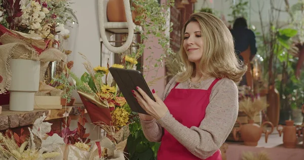 One happy middle aged female entrepreneur holding tablet standing inside flower shop checking for online orders with digital modern device. Small business owner checking inventory managing small business