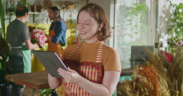 Happy female staff of flower shop holding tablet checking online orders. Young woman employee using modern technology to manage shipment delivery of small business store
