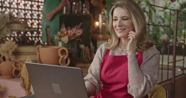 Female entrepreneur owner of flower store taking online orders on the phone in front of laptop computer. Businesswoman talking on cellphone with client