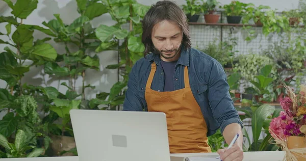 Young florist entrepreneur checking online orders smiling in front of laptop. Small business online orders concept. Male employee wearing apron writing down online orders