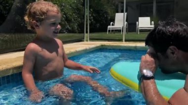 Çocuk ve baba havuzda oynuyorlar, dalga geçiyorlar, su sıçratıyorlar.