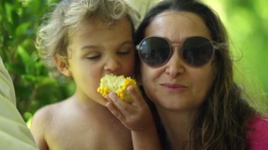 Anne ve çocuk fotoğraf için poz veriyor, anne oğlunu yanağından öpüyor, çocuk mısır yiyor.