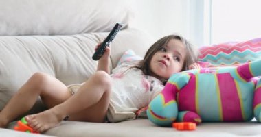 Küçük kız elinde uzaktan kumandayla televizyon izliyor.