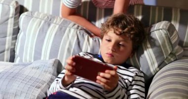 Evde koltukta oturan cep telefonu cihazıyla video oyunu oynayan genç bir çocuk.