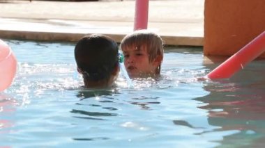 Çocuklar havuz suyunda oynuyorlar, genç çocuk arkadaşa barış işareti yapıyor.