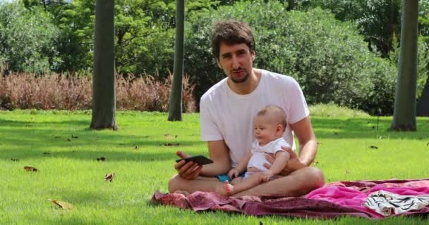 お父さんと赤ちゃんの男の子は草の上に座って自然の中で外の携帯電話のデバイスでコンテンツを見て — ストック動画