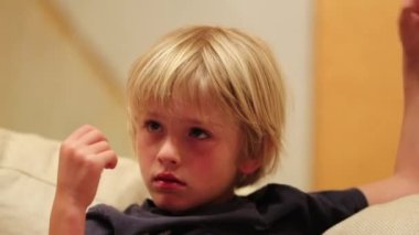 Saçla oynarken televizyon izleyen küçük sarışın çocuk.