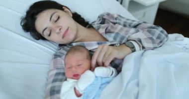 Mother holding newborn baby falling asleep together at hospital after birth in bed