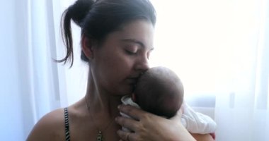 Mother holding newborn baby next to home window, first week of life