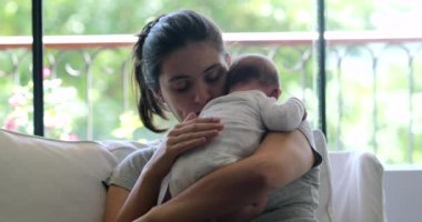 Mother holding newborn baby seated at home sofa tapping at infant back