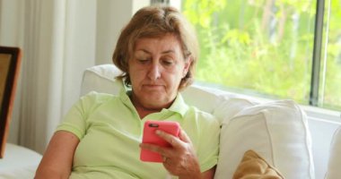 Thoughtful candid older senior woman holding smartphone device seated at home sofa