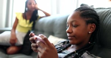 Black girl playing game on phone at home couch, sister watching sibling use smartphone device