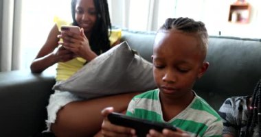 Casual African family relaxing at home sofa using technology. Black parents and kids each using their tech devices bubbles