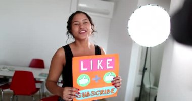 Millennial girl vlogger talking to camera showing sign for audience to like and subscribe to media channel