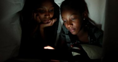 İki kız kardeş gece battaniyenin altında akıllı telefondan video izliyorlar.