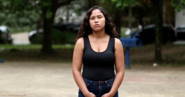 Thoughtful serious hispanic young woman standing outside staring at camera. Portrait close-up face, real people series