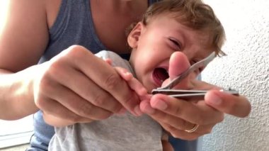 Tearful baby crying while mother trims hand nails. Parent trimming toddler nail
