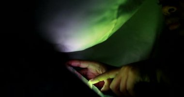 Child hands touching smartphone screen with finger at night under blanket