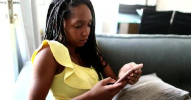 Evdeki kanepede cep telefonu kullanan siyah bir genç kız. Sosyal medya akıllı telefonunda Afrikalı genç kız