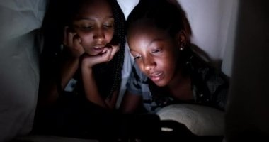Two African teen girls looking at smartphone screen at night under blanket, real life laugh and smile reaction to video content