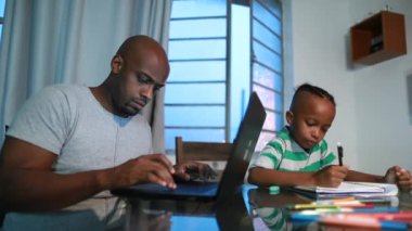 Black African father working from home front laptop. Son doing his homework next to parent