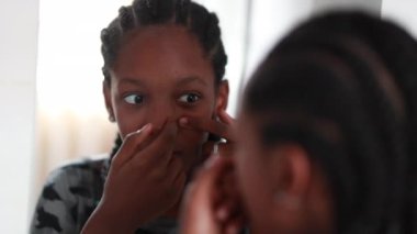 Teen black girl inspecting face in front of mirror. Girl removing zit, pressing with hand