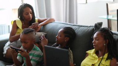 Black African American family sitting at home each in their own bubble using smartphone devices and computer laptop
