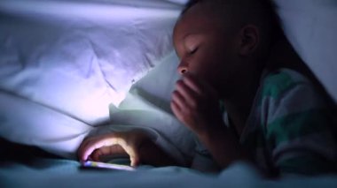 Mixed race child watching content online on cellphone screen under bedroom blanket at night