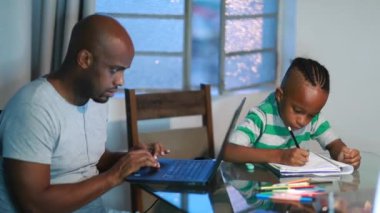 Black family at home, son doing homework, father working in front of laptop computer