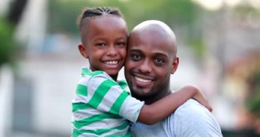 Black father and son smiling at camera. Child hugging dad outdoors, looking at camera