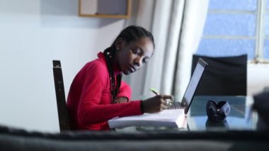 African girl taking notes studying at home in front of laptop computer